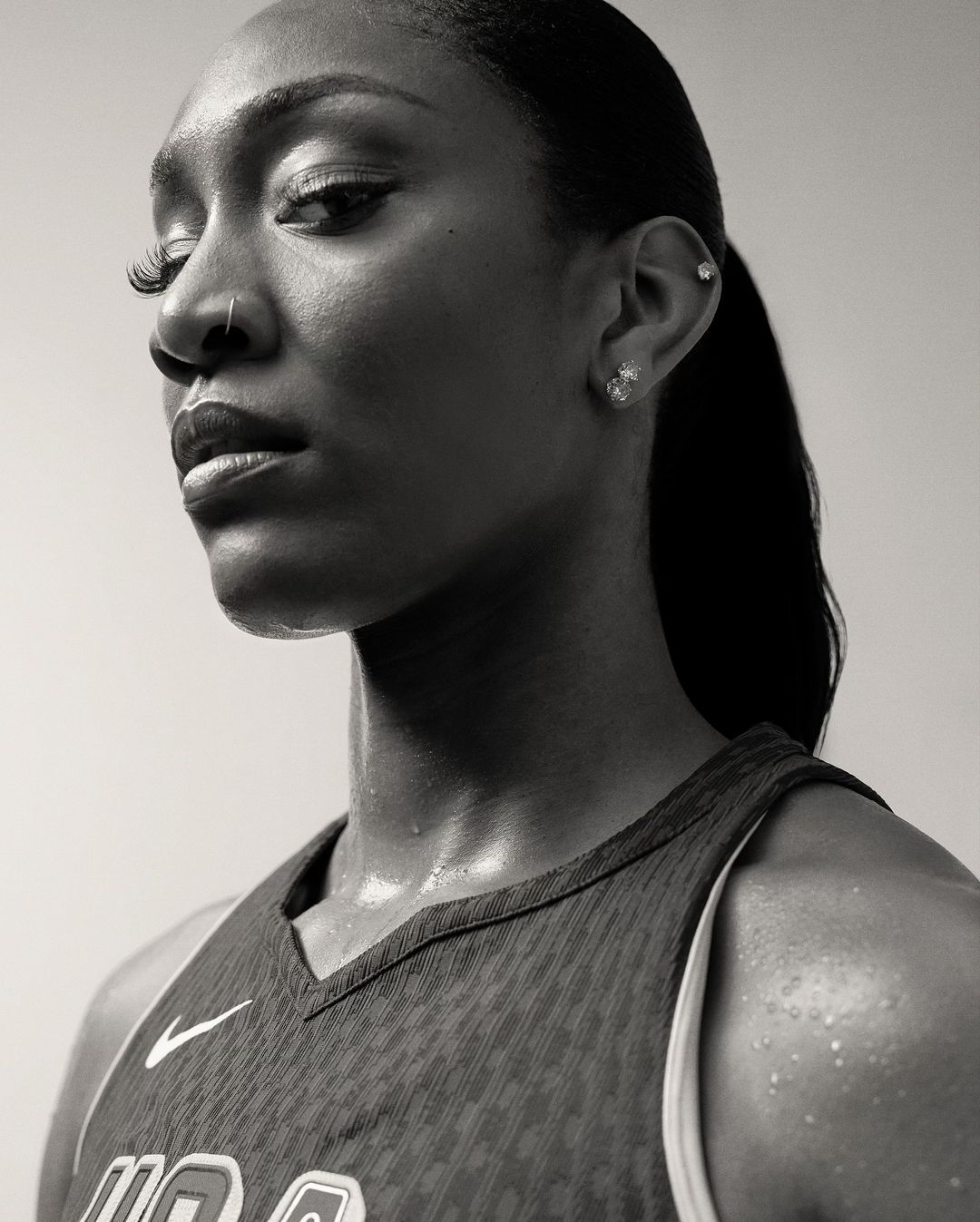Monochrome portrait of a Black woman wearing a sports jersey with visible sweat, looking off camera with neutral expression against plain backdrop.