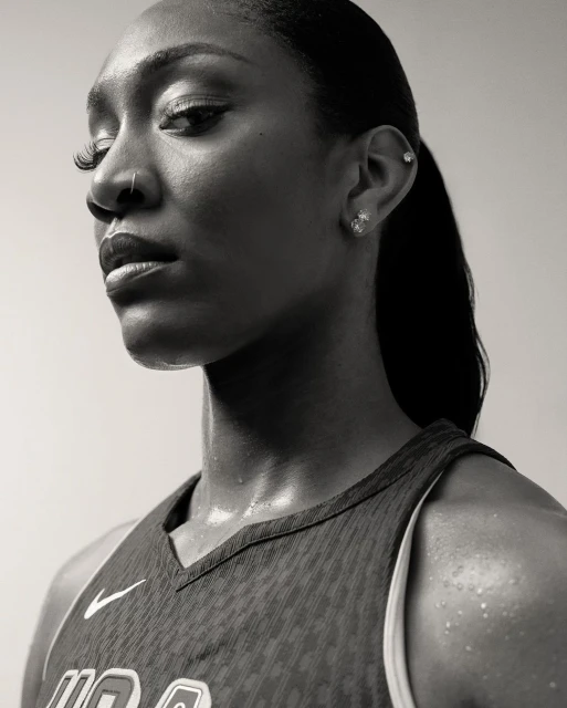 Monochrome portrait of a Black woman wearing a sports jersey with visible sweat, looking off camera with neutral expression against plain backdrop.