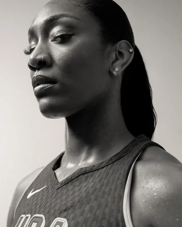 Monochrome portrait of a Black woman wearing a sports jersey with visible sweat, looking off camera with neutral expression against plain backdrop.