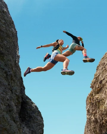 Two athletes leap between rock formations against a clear blue sky, showcasing fitness, athleticism, and outdoor running.