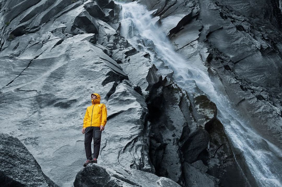 Man in yellow jacket stands on rock formation with mountain and waterfall backdrop, creating a striking outdoor landscape image.
