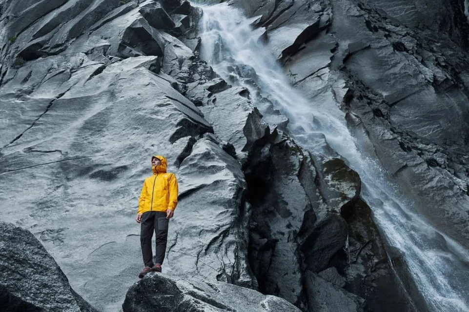 Man in yellow jacket stands on rock formation with mountain and waterfall backdrop, creating a striking outdoor landscape image.