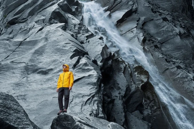 Man in yellow jacket stands on rock formation with mountain and waterfall backdrop, creating a striking outdoor landscape image.