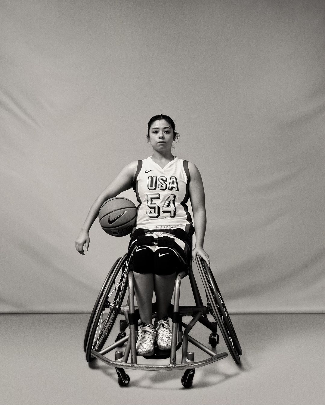 Black and white portrait of a woman in a USA basketball jersey seated in a sports wheelchair, holding a basketball, indoor setting.