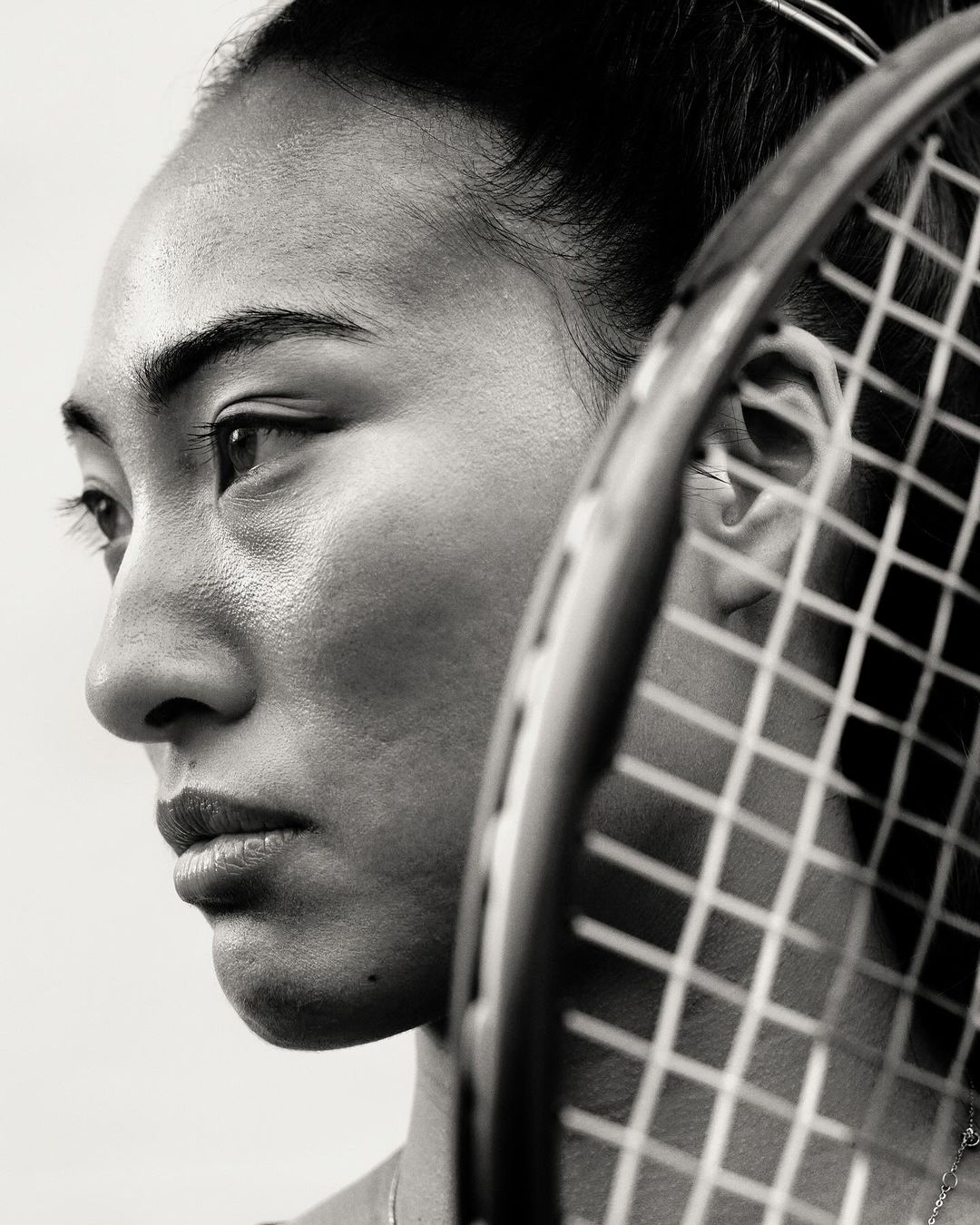 Close-up black and white portrait of an Asian woman in profile with a tennis racket obscuring part of her face.