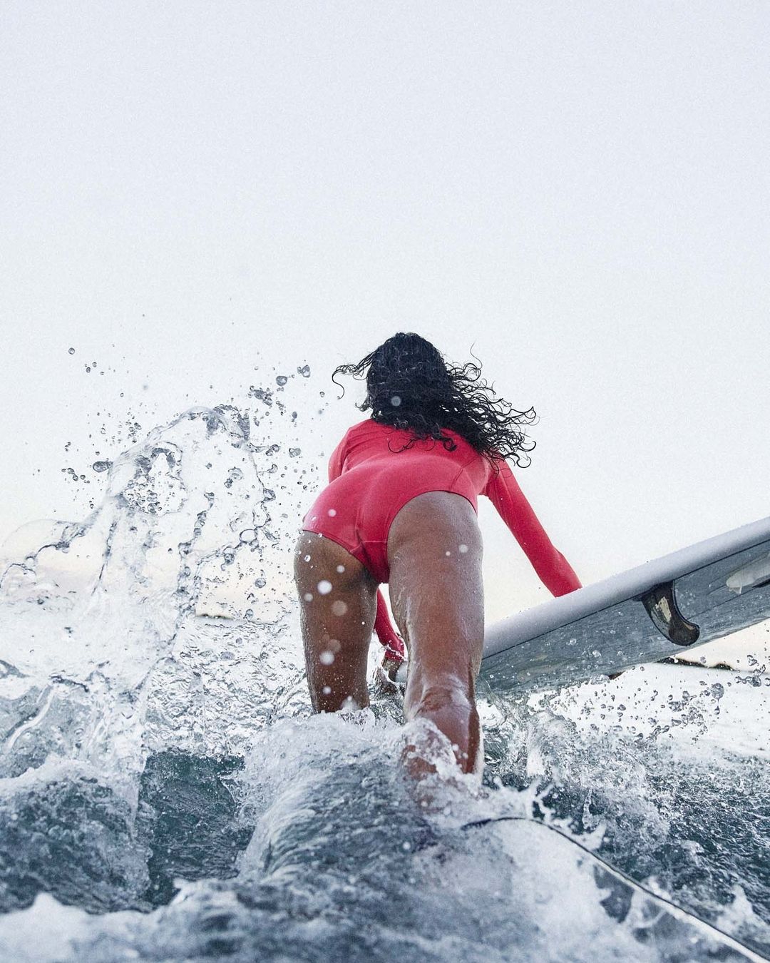 Woman in a pink wetsuit is surfing on a wave with a surfboard. The shot is from behind with the focus on the surfer.