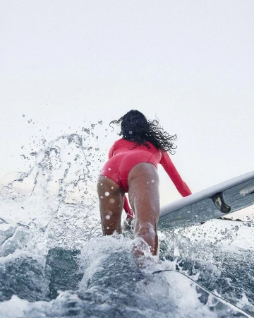 Woman in a pink wetsuit is surfing on a wave with a surfboard. The shot is from behind with the focus on the surfer.