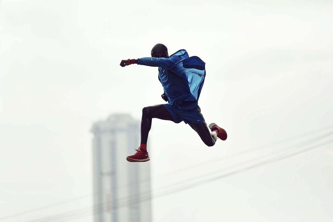 Adult male athlete in blue sportswear and red sneakers mid-jump against a soft white background with blurred cityscape.