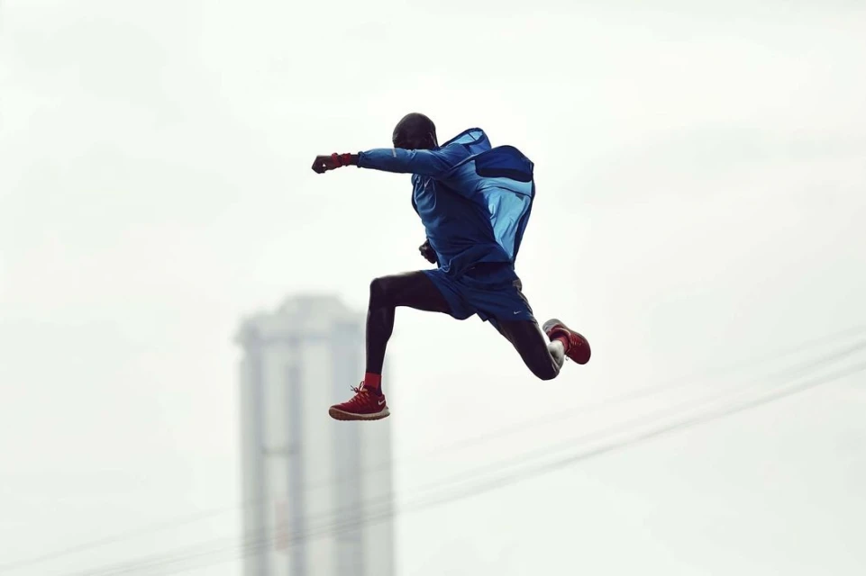 Adult male athlete in blue sportswear and red sneakers mid-jump against a soft white background with blurred cityscape.