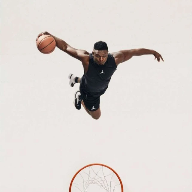 An athletic Black male in sportswear dynamically jumps with a basketball against a bright background, captured from a low angle, hoop visible.