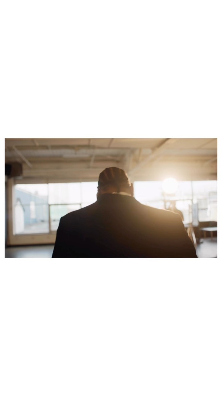 A man in a dark suit seen from behind standing in an interior space with bright window backlighting.