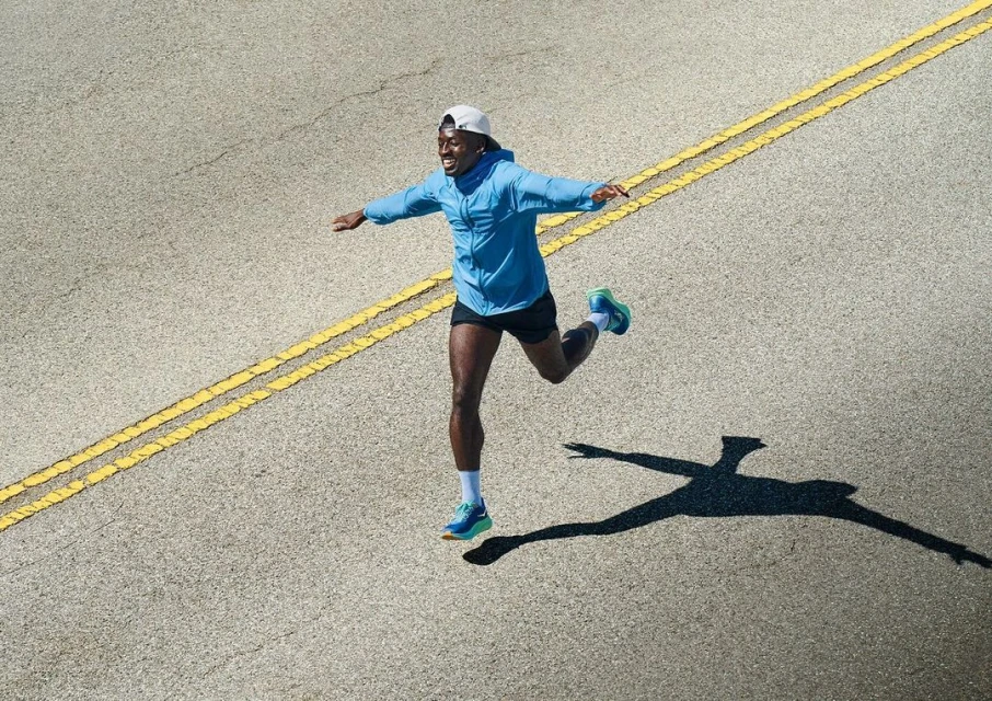 Athletic man in blue jacket and shorts runs on asphalt road with yellow lines in bright daylight, casting a long shadow. 