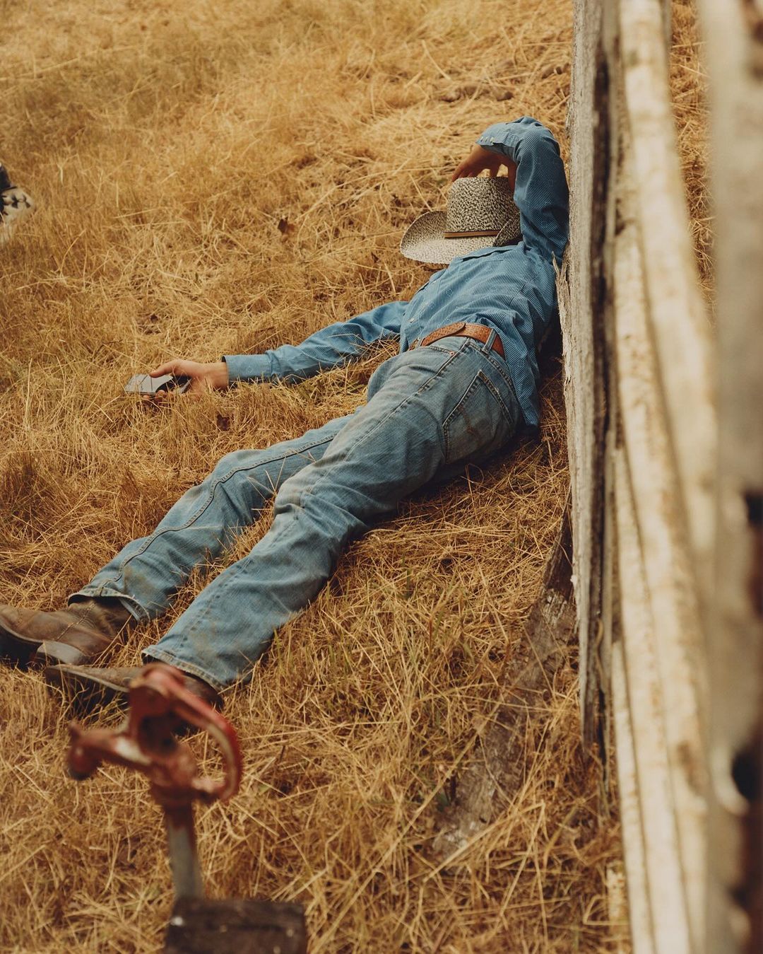 Man in denim shirt and jeans lying on grass with hat covering face next to fence, outdoor setting, natural light, and neutral tones.