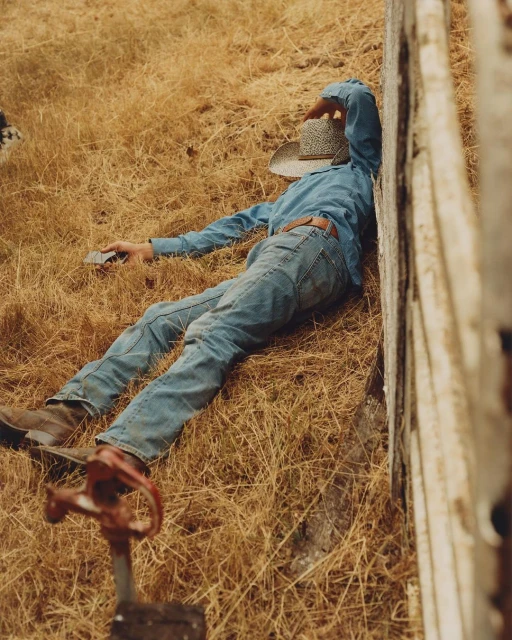 Man in denim shirt and jeans lying on grass with hat covering face next to fence, outdoor setting, natural light, and neutral tones.
