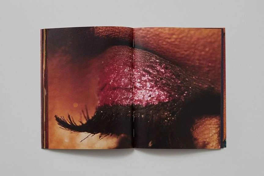 Macro shot in an open book reveals a human eye with striking pink glitter makeup on the eyelid and defined eyelashes.