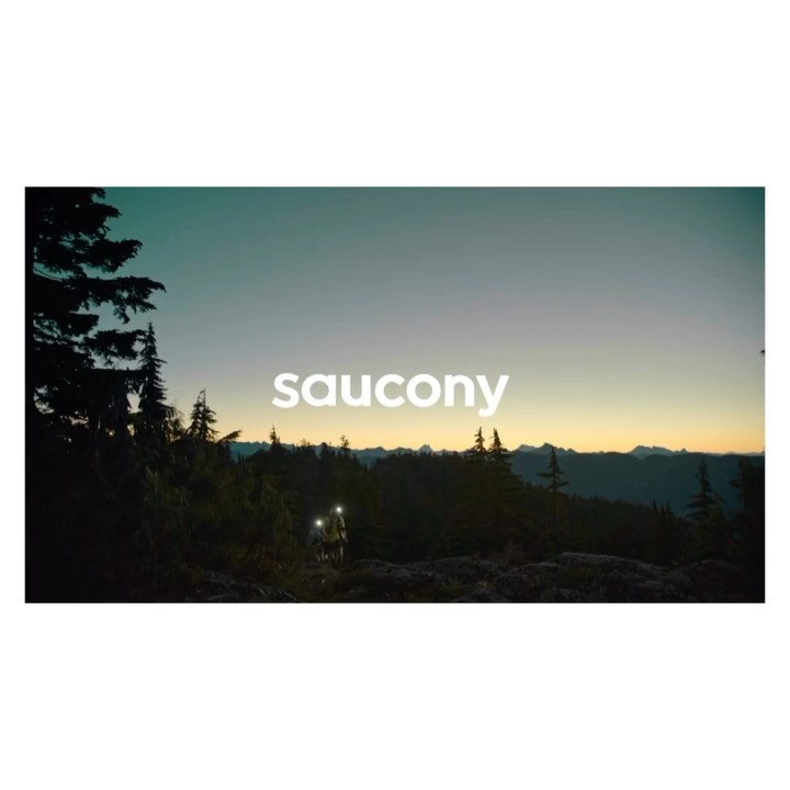 Two people hike through a dark forest at dusk, illuminated by headlamps, near silhouetted mountains under a twilight sky. Saucony logo.