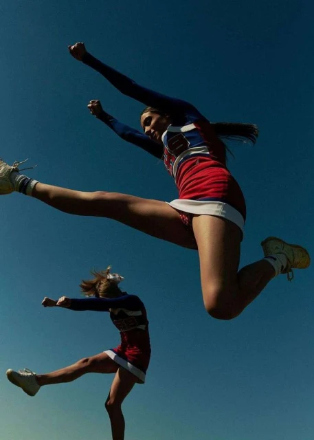 Two female cheerleaders jumping in the air wearing red, white and blue uniforms against a clear blue sky. The cheerleaders are in motion.