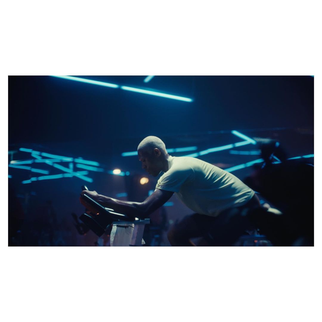 An African American man with a shaved head exercises on a stationary bike in a dimly lit gym featuring blue neon lighting.