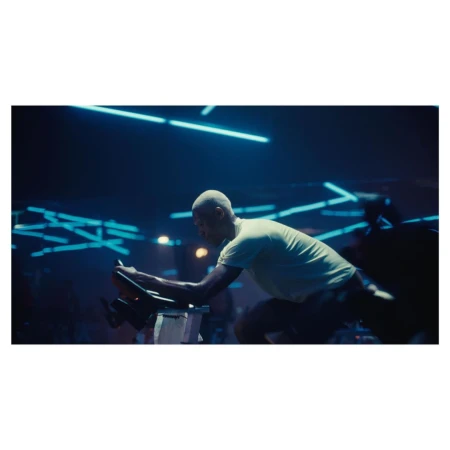 An African American man with a shaved head exercises on a stationary bike in a dimly lit gym featuring blue neon lighting.