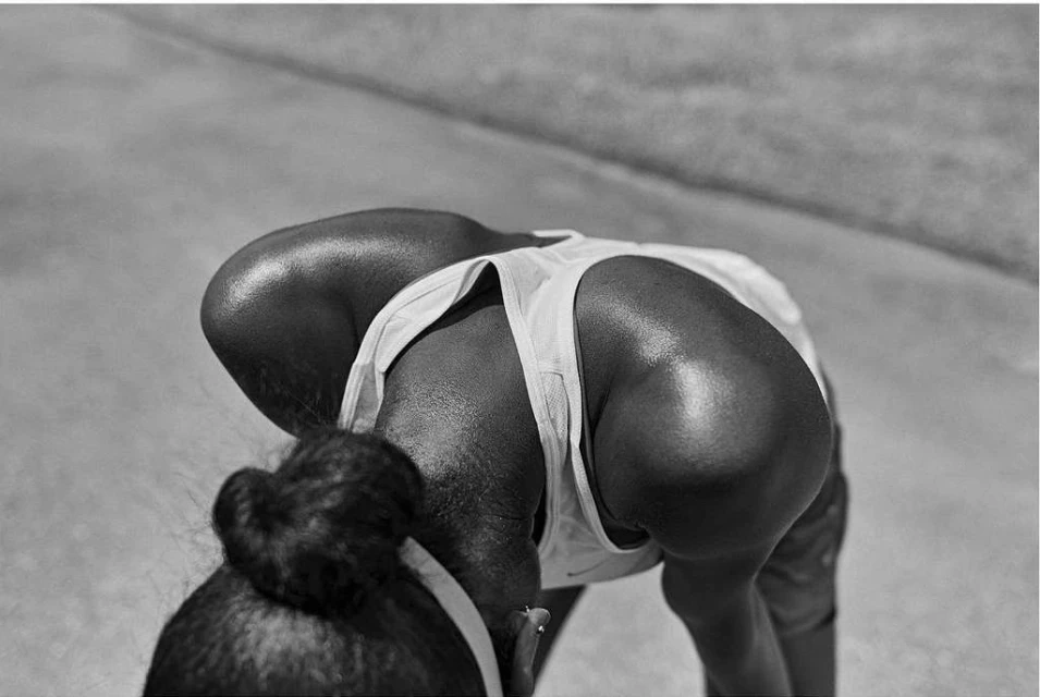 Black and white photo of an adult female athlete in a tank top, bending over with her hair tied back, and shiny skin.