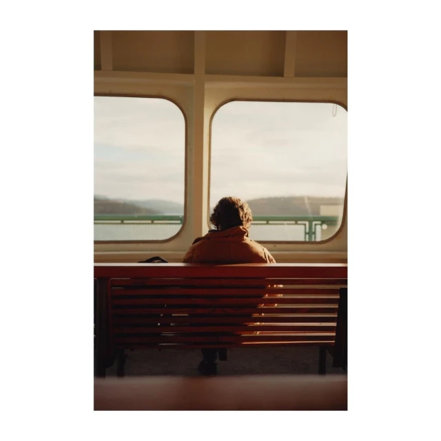 An adult sits on a bench inside a ferry looking out the window at the water and distant land, with the window frame in view.