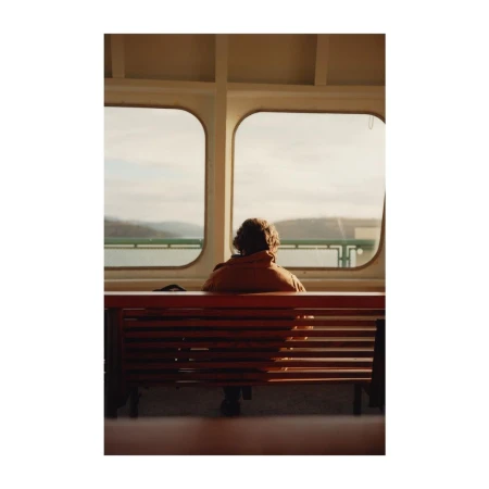 An adult sits on a bench inside a ferry looking out the window at the water and distant land, with the window frame in view.