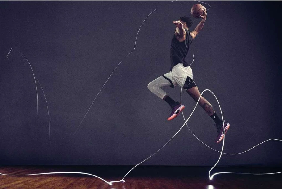 A man wearing sportswear leaps mid-air with a basketball in a studio, highlighted by dynamic white light trails on a wood floor.