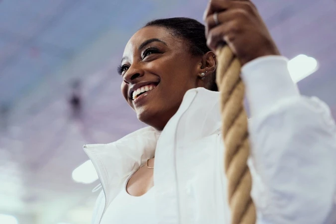 Simone Biles smiling while gripping a thick climbing rope in a gym, wearing a white jacket.