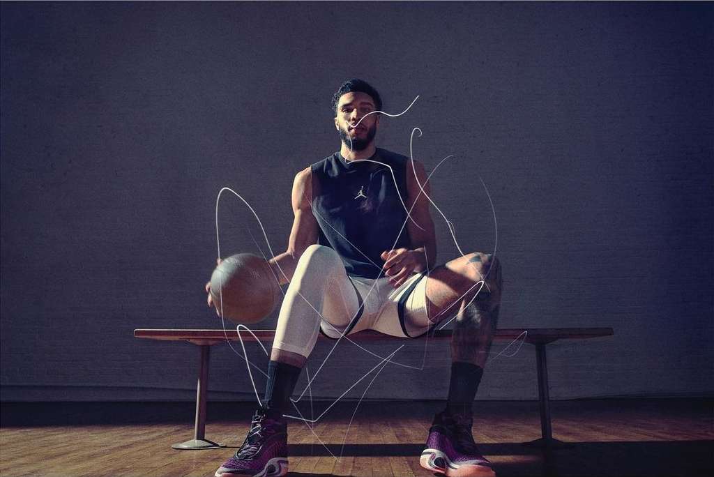 Athlete in tank top sits on bench with basketball, wearing Jordan shoes with artistic light trails and gray background.