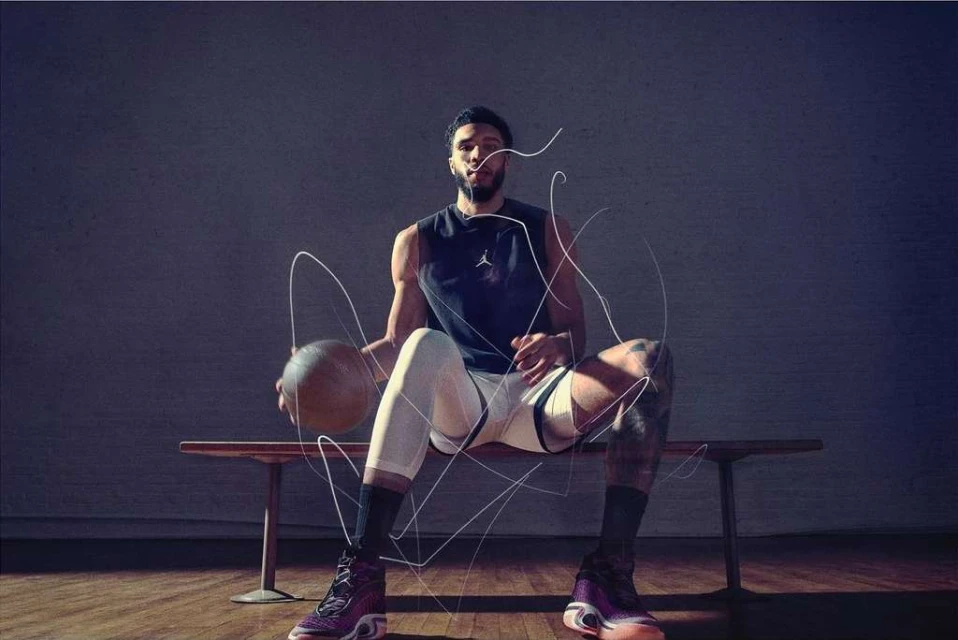Athlete in tank top sits on bench with basketball, wearing Jordan shoes with artistic light trails and gray background.