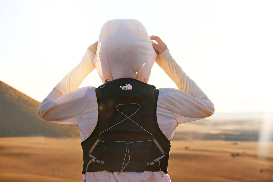 A person stands on a hill wearing a black hydration pack, white hooded shirt, and looking toward the horizon on a bright day.
