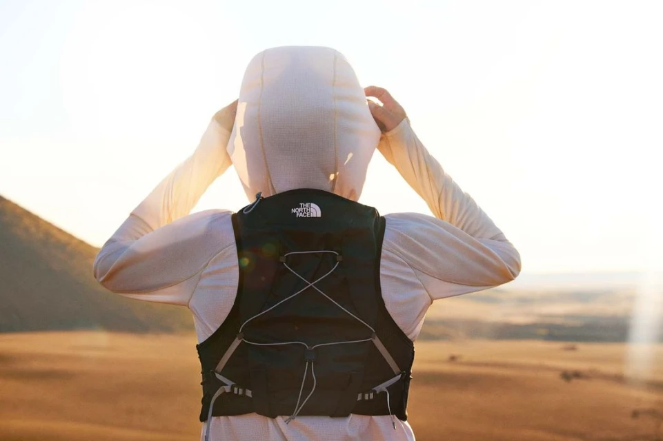 A person stands on a hill wearing a black hydration pack, white hooded shirt, and looking toward the horizon on a bright day.