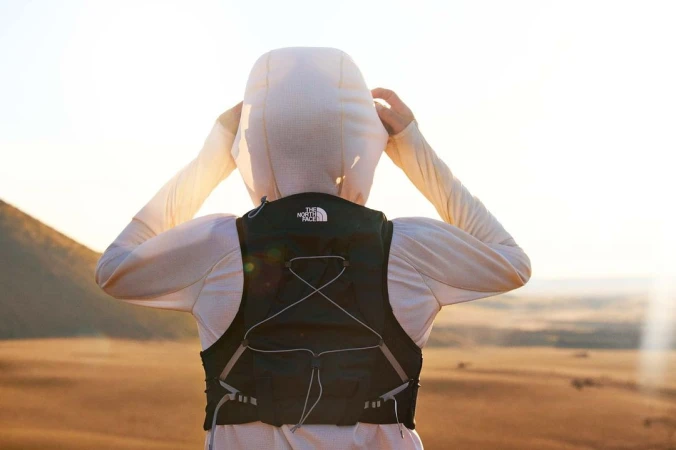 A person stands on a hill wearing a black hydration pack, white hooded shirt, and looking toward the horizon on a bright day.