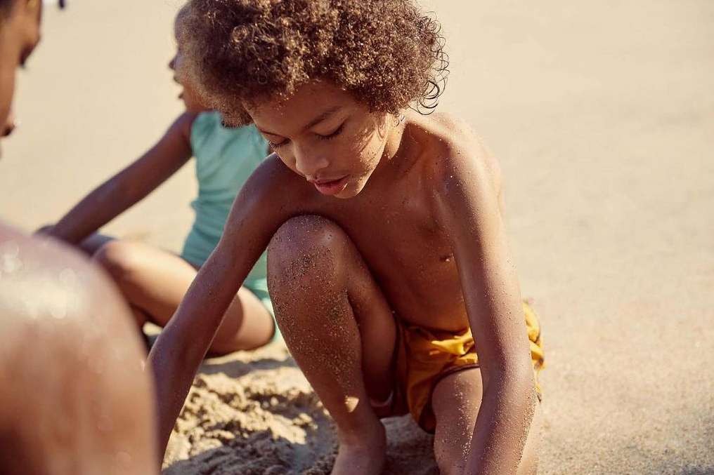 Child plays in sand on a beach, with a partially visible adult and another child in the background enjoying a sunny day by the sea.