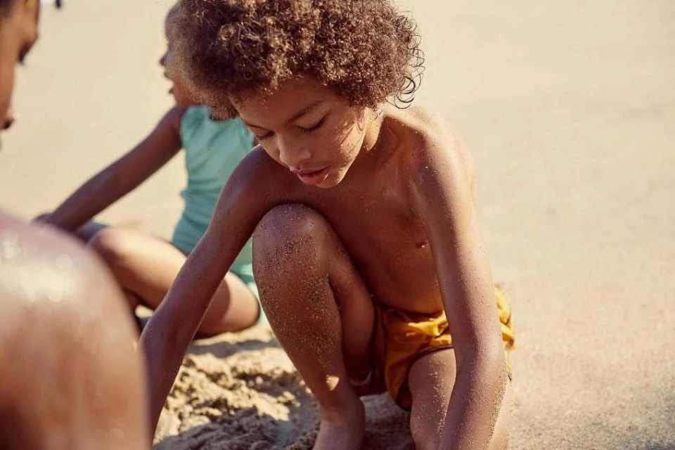 Child plays in sand on a beach, with a partially visible adult and another child in the background enjoying a sunny day by the sea.