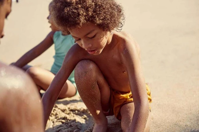 Young child with curly hair crouching in the sand at the beach playing with sand, photographed by Anthony Blasko.