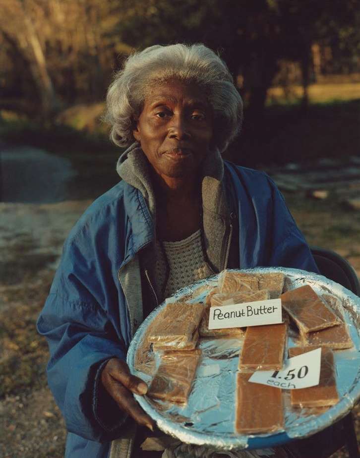 Elderly African-American woman outdoors holding a silver tray with rectangular pieces of peanut butter fudge for sale.