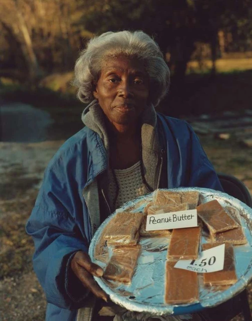 Elderly African-American woman outdoors holding a silver tray with rectangular pieces of peanut butter fudge for sale.