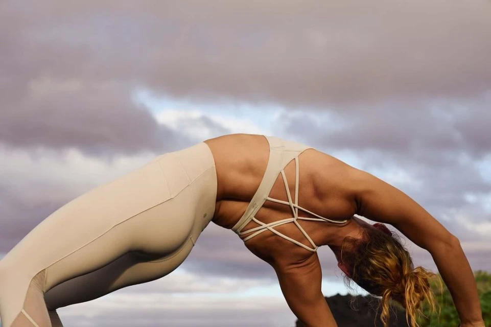 An adult woman practices yoga outdoors with a backbend pose, wearing a light-colored sports bra and leggings against a cloudy sky backdrop.