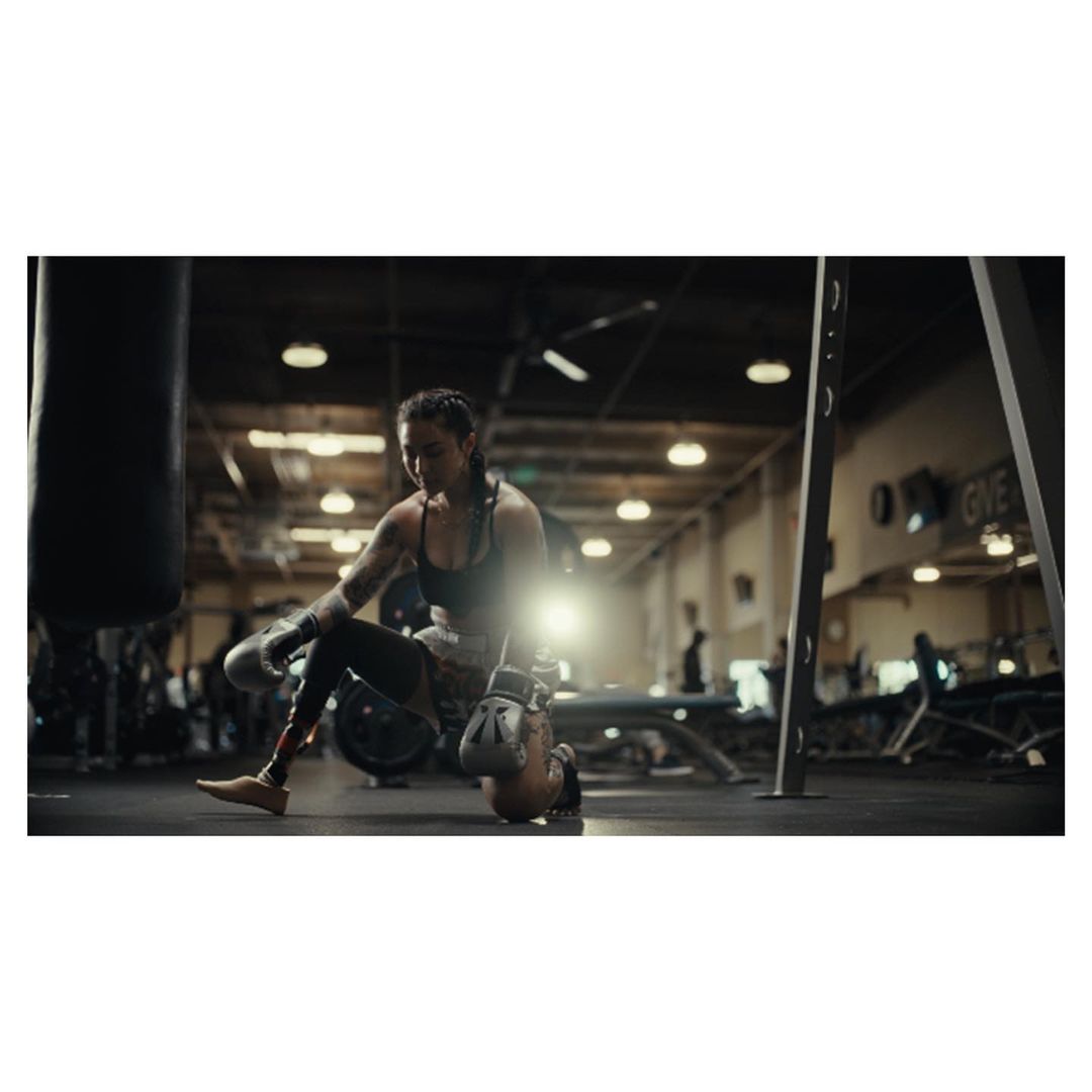 Female boxer on one knee at the gym with boxing gloves next to a punching bag, wearing athletic apparel, showcasing fitness lifestyle.