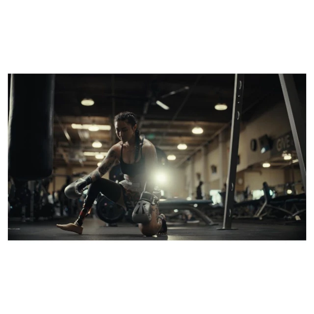 Female boxer on one knee at the gym with boxing gloves next to a punching bag, wearing athletic apparel, showcasing fitness lifestyle.