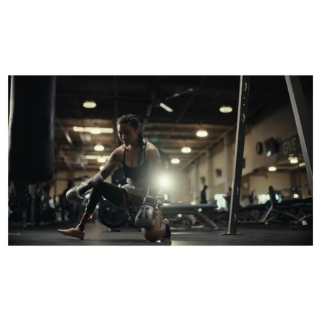 Female boxer on one knee at the gym with boxing gloves next to a punching bag, wearing athletic apparel, showcasing fitness lifestyle.
