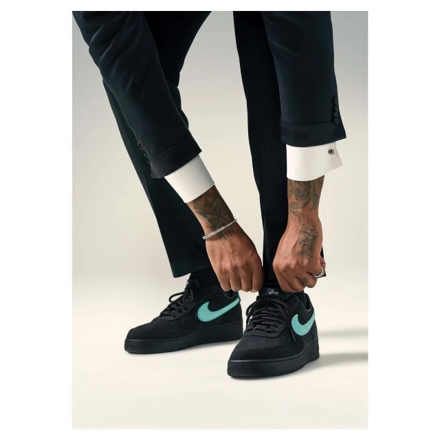Close up of black sneakers with tiffany blue swoosh logo, laced by a man in a dark suit against a light neutral backdrop.