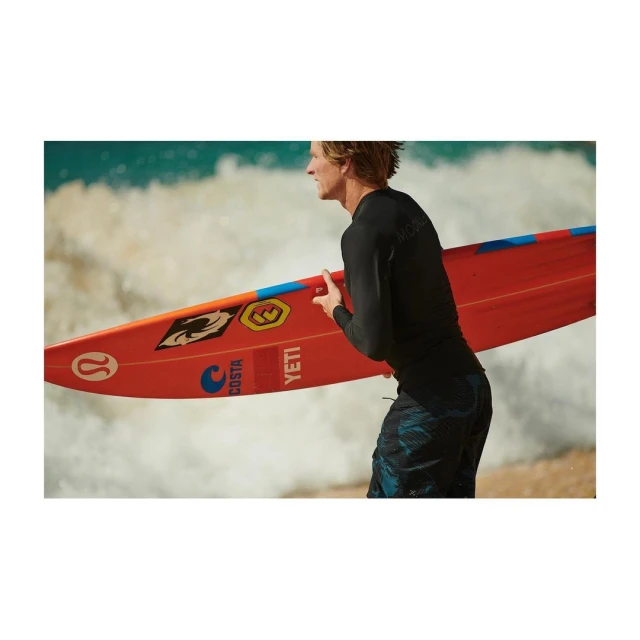 A male surfer carries a bright orange surfboard on a sandy beach with ocean waves crashing in the background on a sunny day.