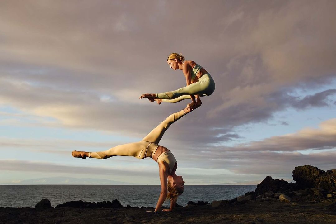 Two women performing acroyoga on a rocky beach at sunset. The woman on the bottom is balancing the other woman on her feet.