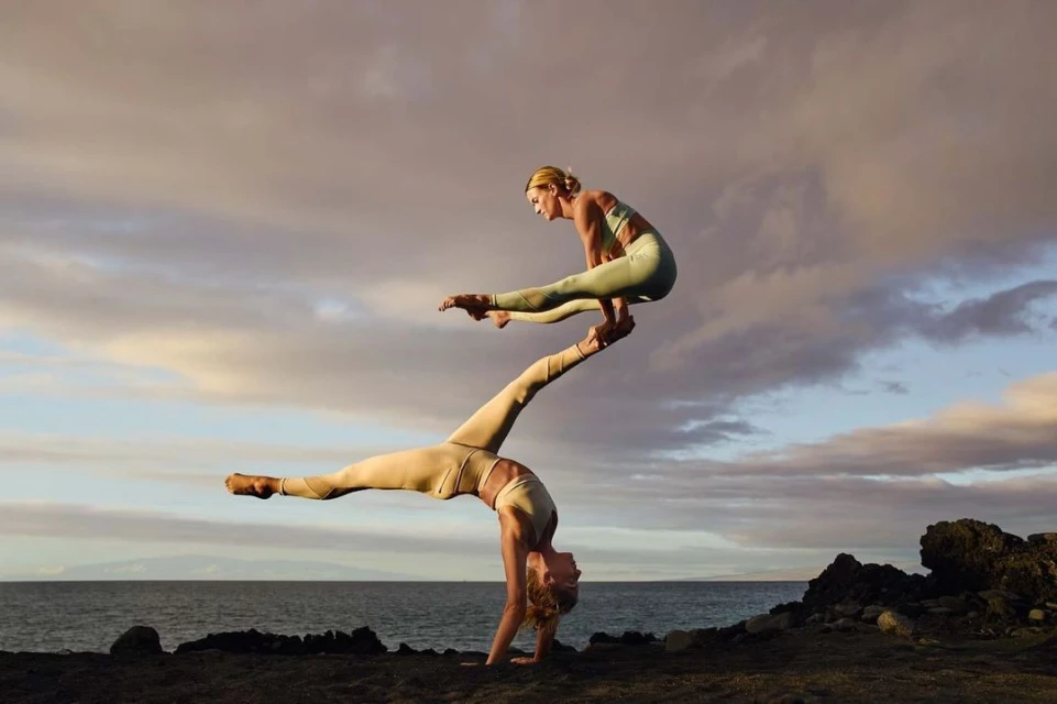 Two women performing acroyoga on a rocky beach at sunset. The woman on the bottom is balancing the other woman on her feet.