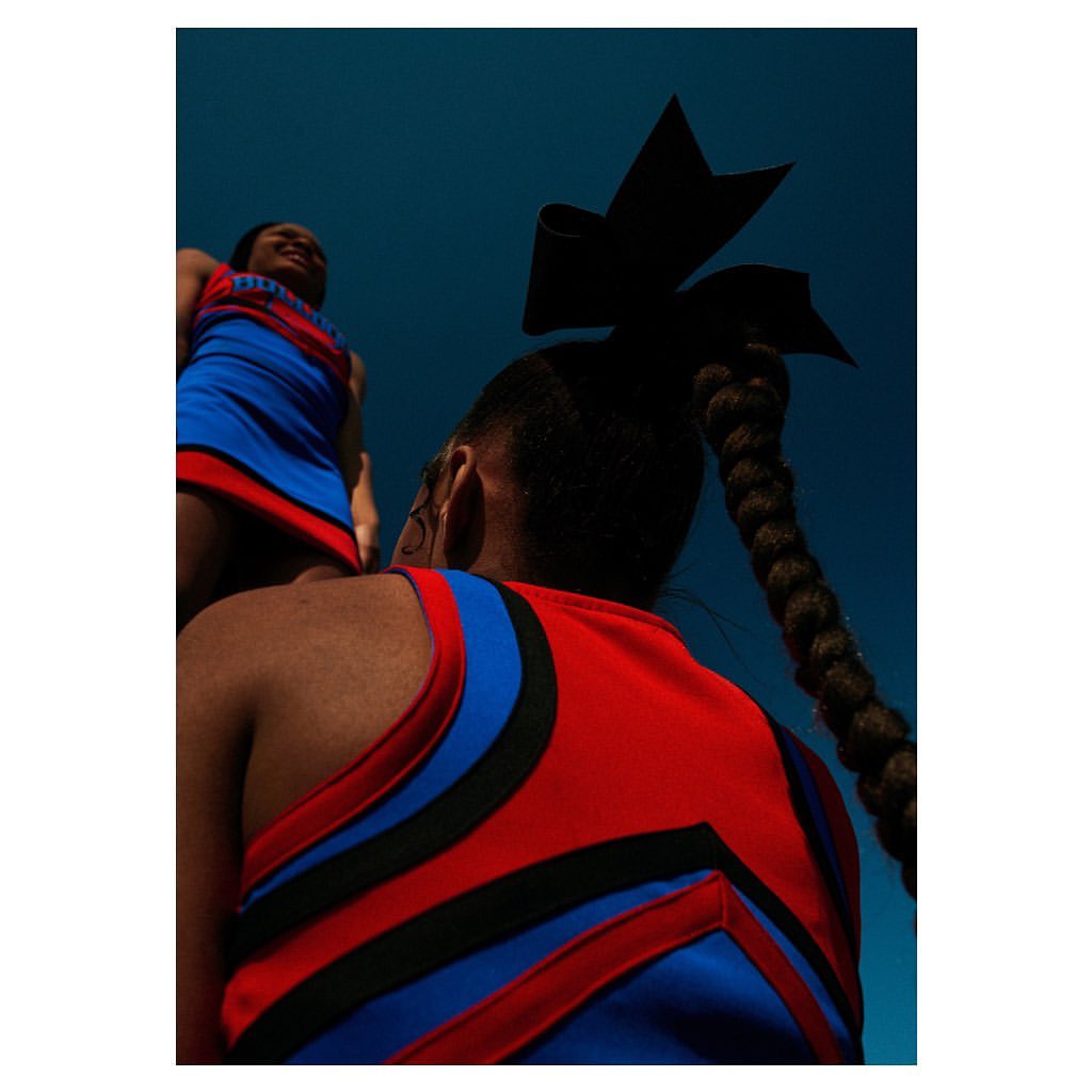 Two cheerleaders in red and blue uniforms pose against a blue sky, one with a bow and braid, shot from a low angle looking up.