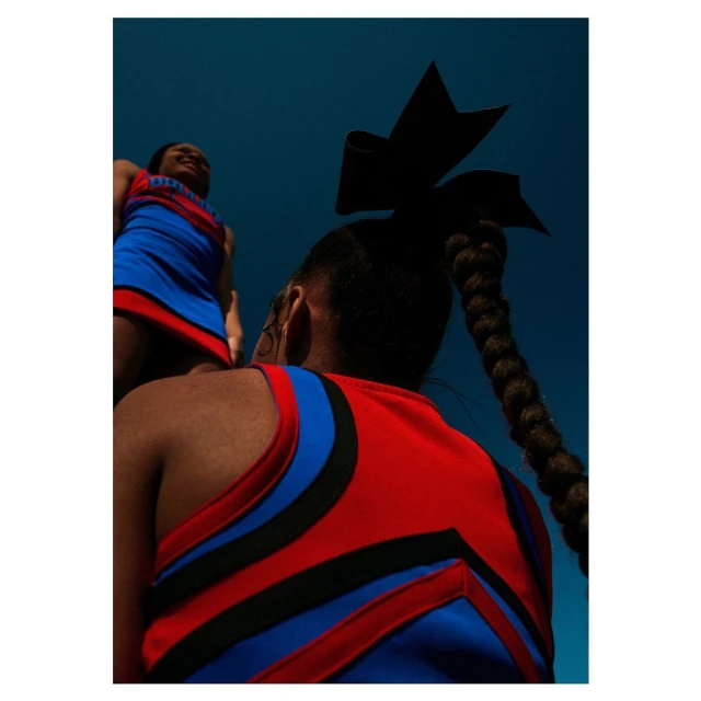 Two cheerleaders in red and blue uniforms pose against a blue sky, one with a bow and braid, shot from a low angle looking up.