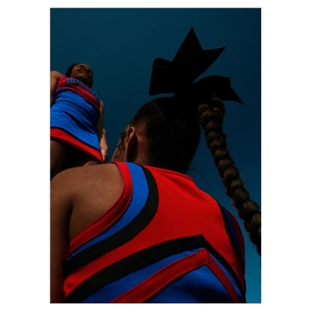 Two cheerleaders in red and blue uniforms pose against a blue sky, one with a bow and braid, shot from a low angle looking up.