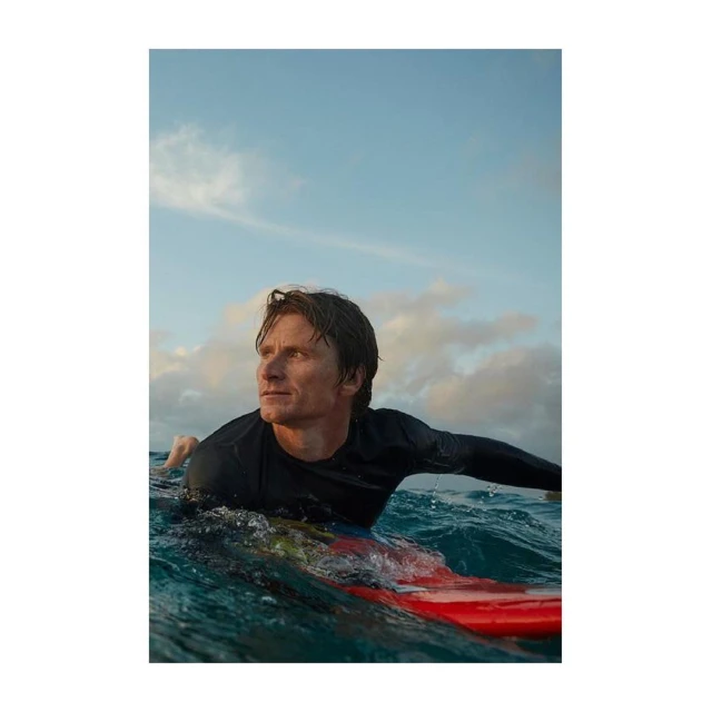 Man paddles on a red surfboard through ocean water, looking towards the horizon, with a bright blue sky and scattered clouds above.